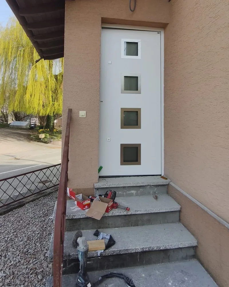 Porte d’entrée blanche à quatre petites fenêtres carrées, le jour de la pose, outils encore sur les marches — photo de chantier
